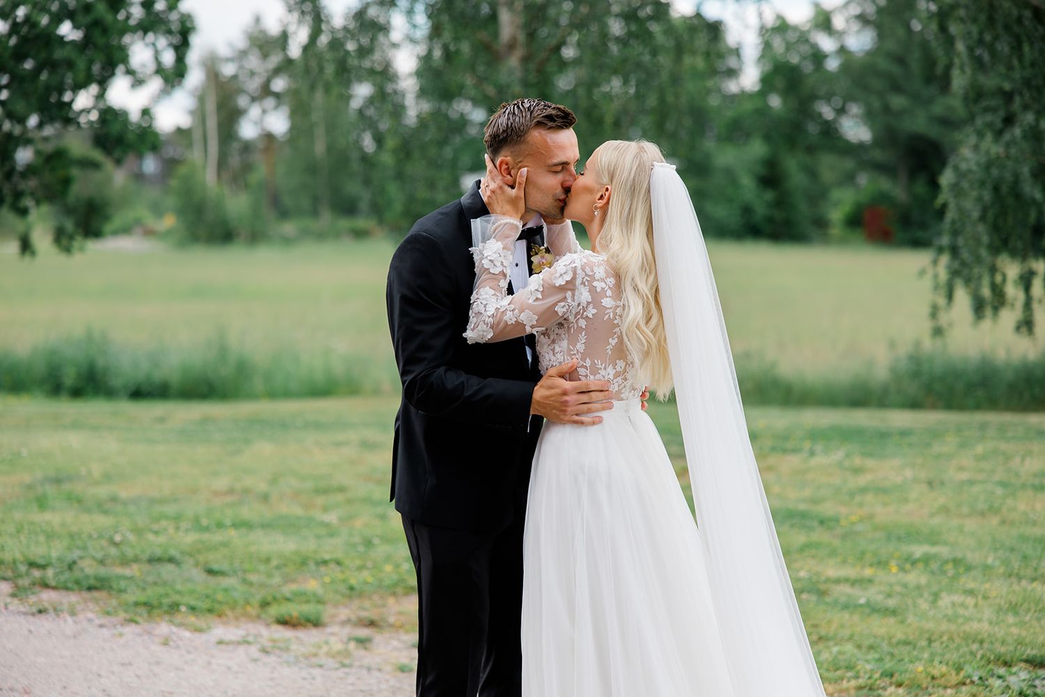 A couple shares an intimate moment in their wedding attire on a path with green trees and fields in the background.