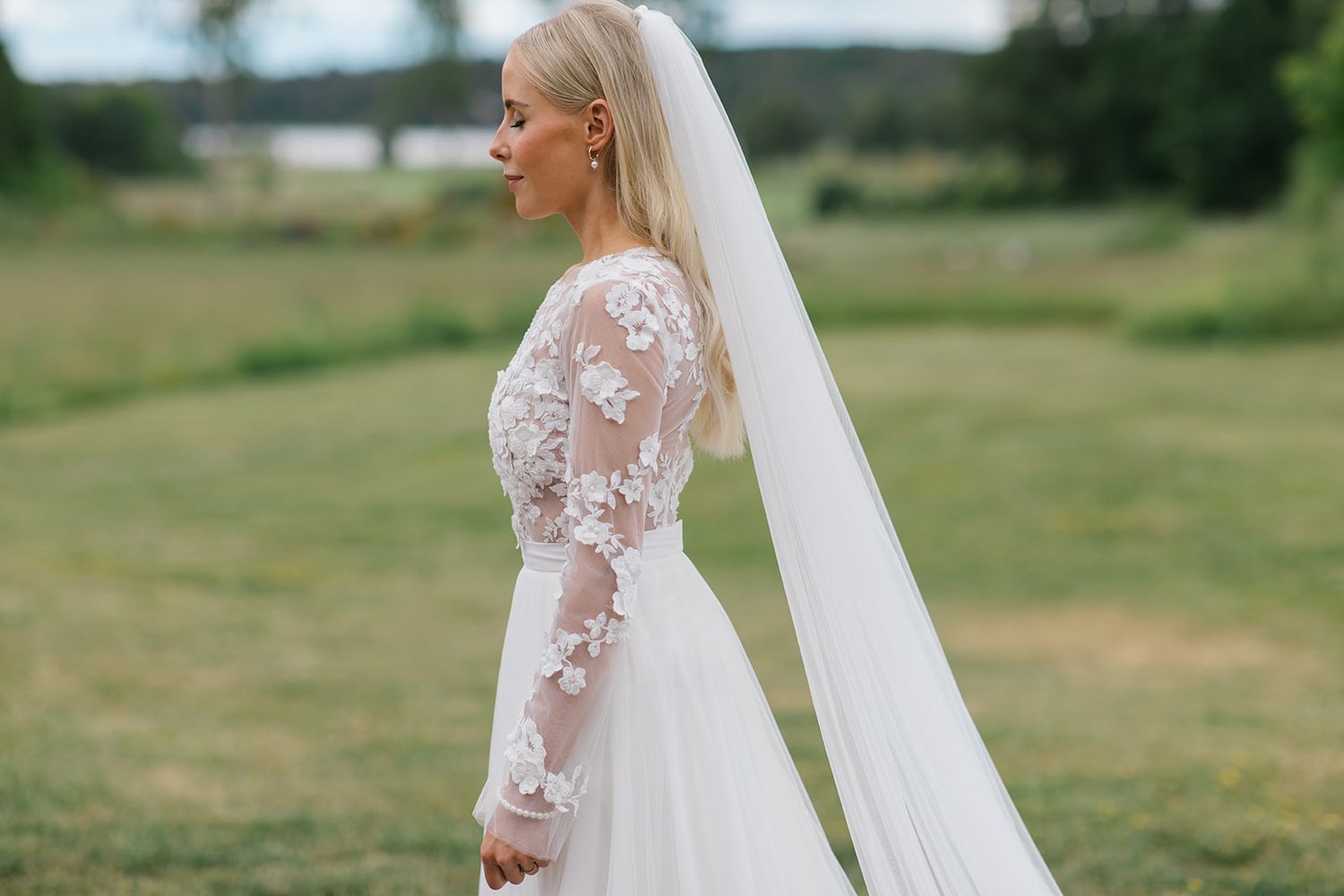 A bride in a long-sleeved lace wedding gown with flowing veil stands in profile against a green countryside backdrop.