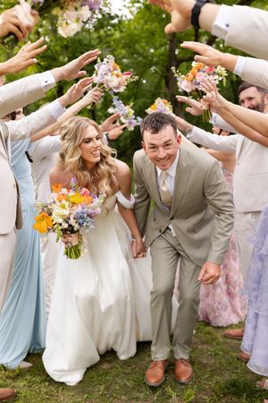 Wedding party celebrates with flower petals as couple walks under raised arms during outdoor garden ceremony.