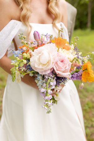 A colorful spring wedding bouquet with roses, poppies and wildflowers held against a white wedding dress outdoors.