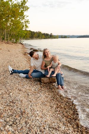 A couple relaxes together on a rocky beach shoreline at sunset with water and trees in the background.