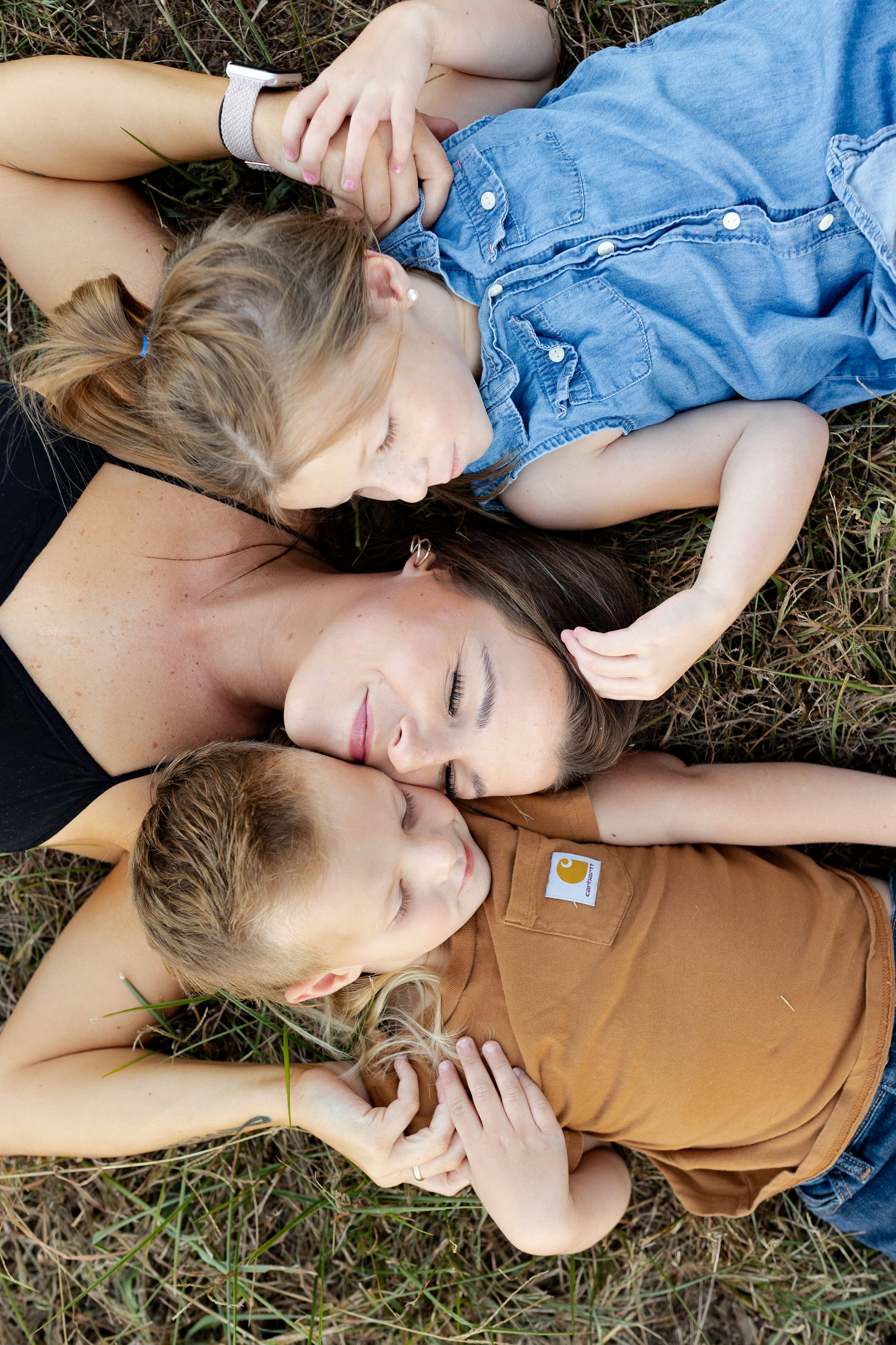 Three people lying on grass in a circle formation wearing casual summer clothing.