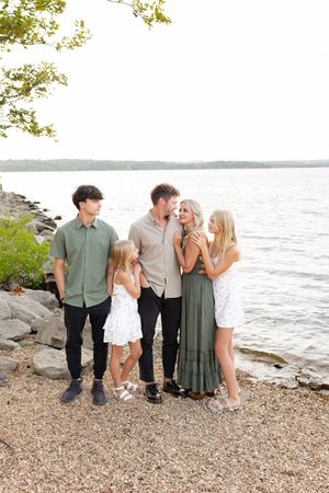 Family group poses together on a rocky beach shoreline at sunset with water and trees in background.