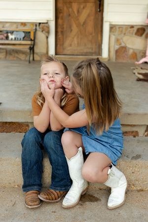 Two children in casual clothes sitting together on concrete steps near a wooden door.