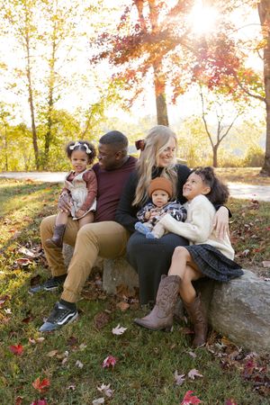 A family sits together on a large rock surrounded by fallen autumn leaves during a golden sunset photoshoot.