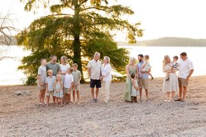Group in coordinated light colored outfits gathered on a beach shoreline at sunset with a large tree and lake view.