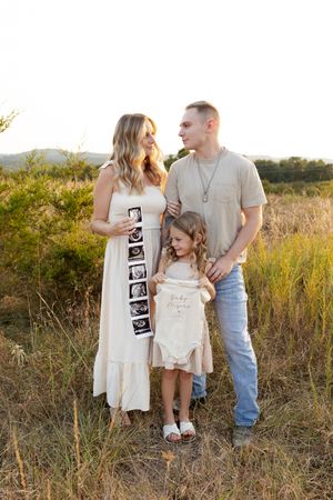 Family stands together in a golden field during sunset holding pregnancy announcement signs and ultrasound photos.