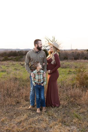 A couple and child stand together in a rustic autumn field at sunset wearing casual attire.