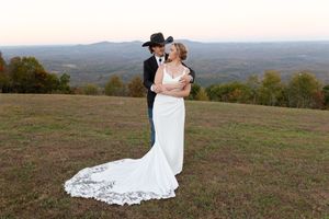 A couple in wedding attire embraces while standing in a scenic mountain meadow with rolling hills in the background.