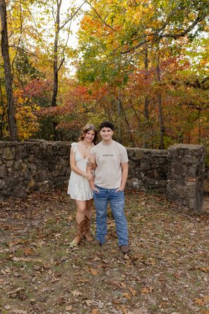 A couple stands together against a stone wall surrounded by vibrant fall foliage in a forest setting.