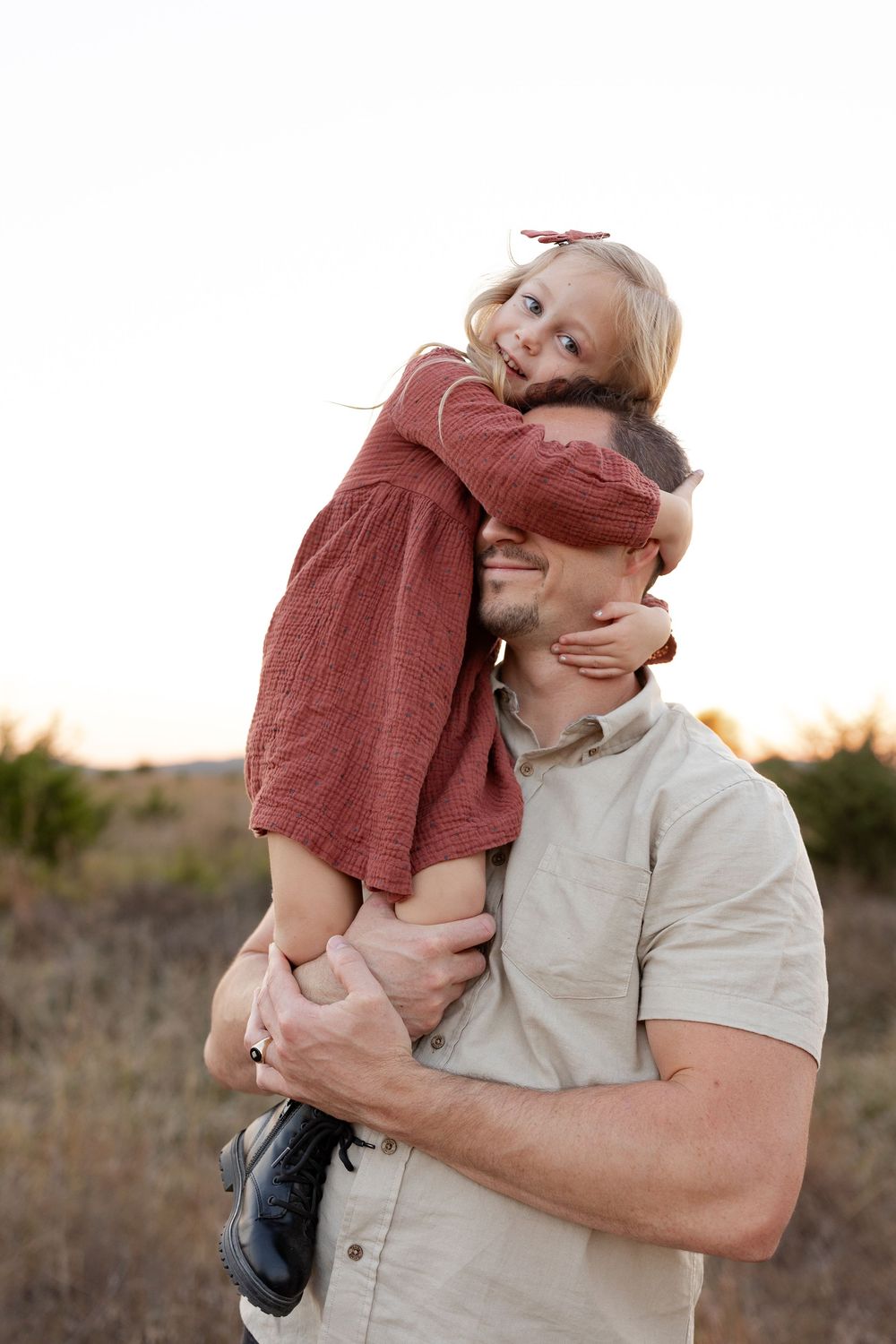 A parent in a beige shirt embraces and lifts a small child wearing red in an outdoor setting at sunset.