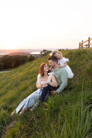 A family sits together on a grassy hillside at sunset overlooking a scenic mountain landscape.