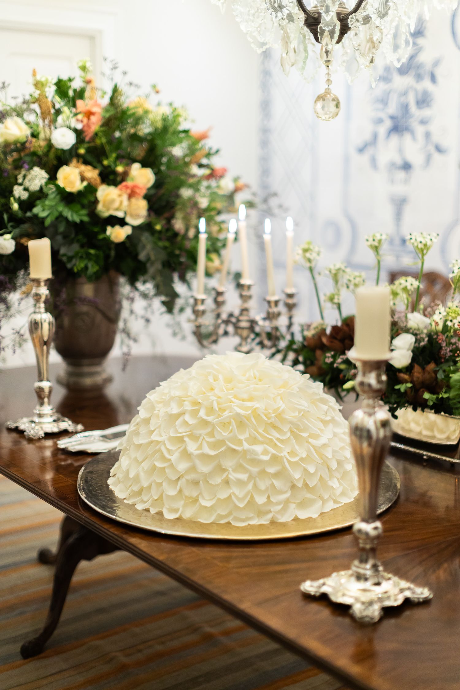 Elegant white ruffled wedding cake displayed on a wooden table with floral arrangements and candles in the background.