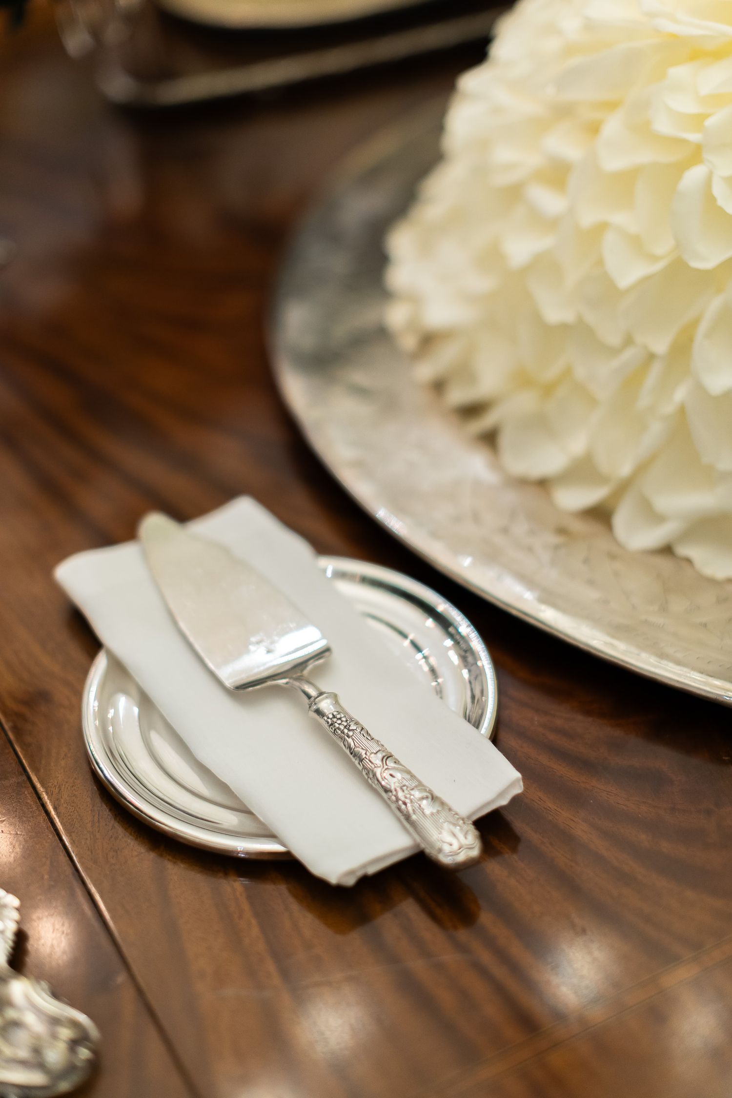 Vintage silverware with ornate handles lies beside white plates on a dark wooden table surface.