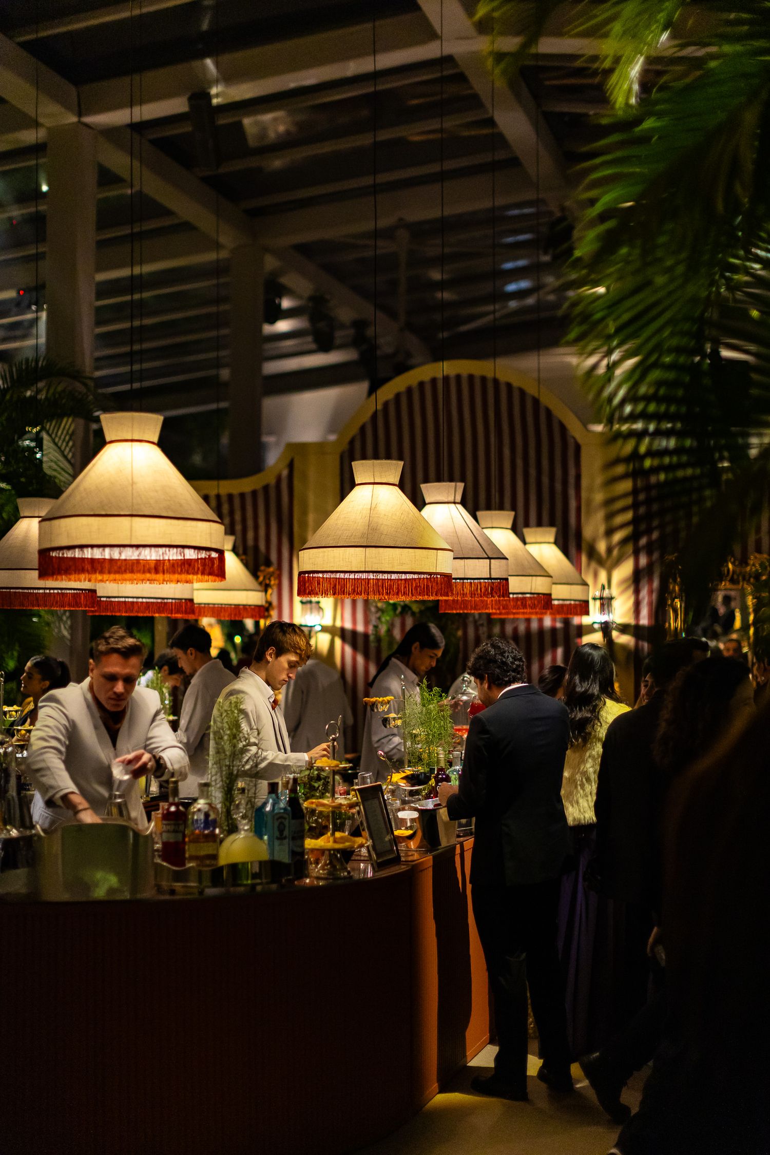 People gather at a dimly lit restaurant bar with hanging pendant lamps and tropical plants creating an elegant ambiance.