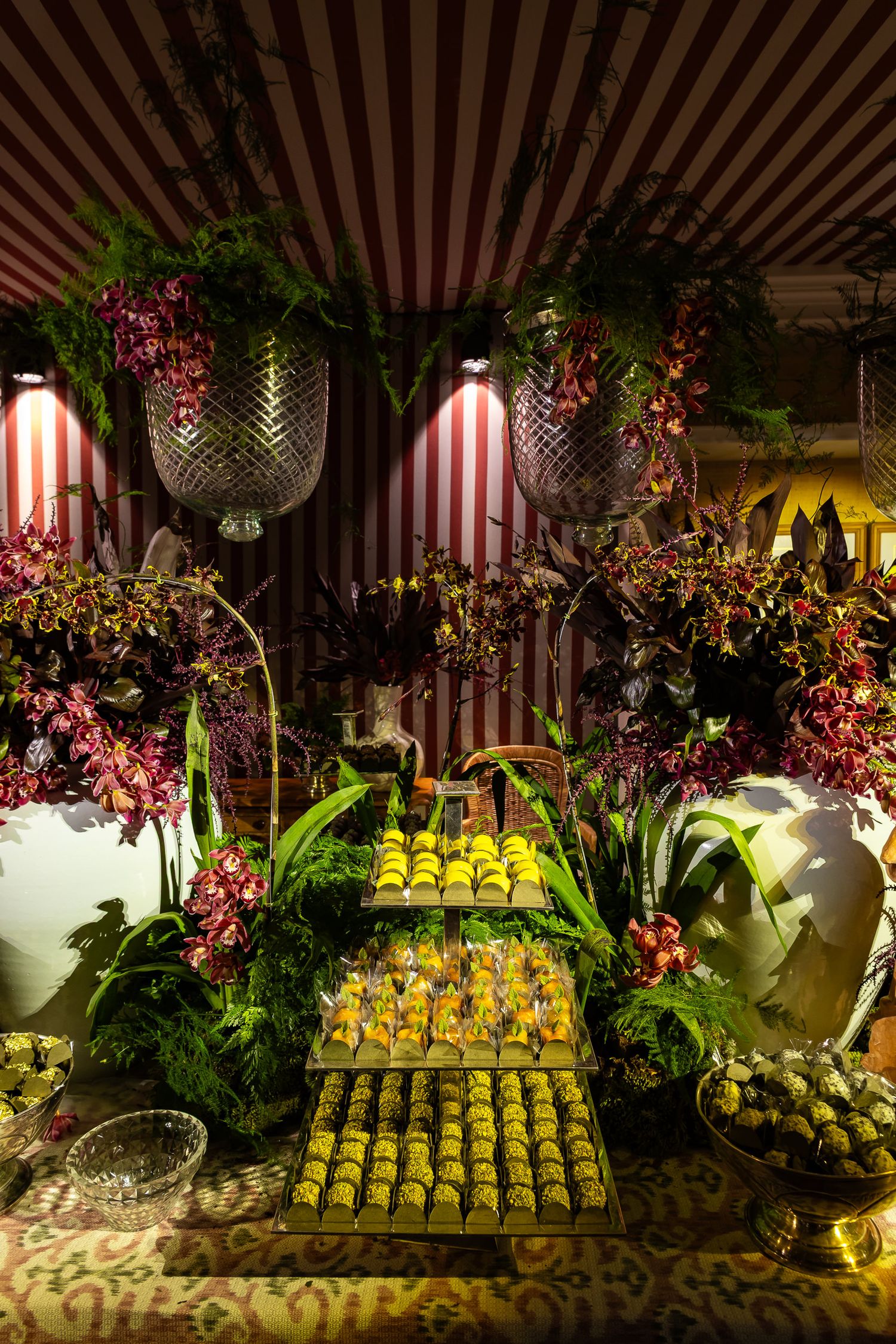 Elegant dessert display with yellow macarons arranged on tiered stands amid floral decorations and striped backdrop lighting.