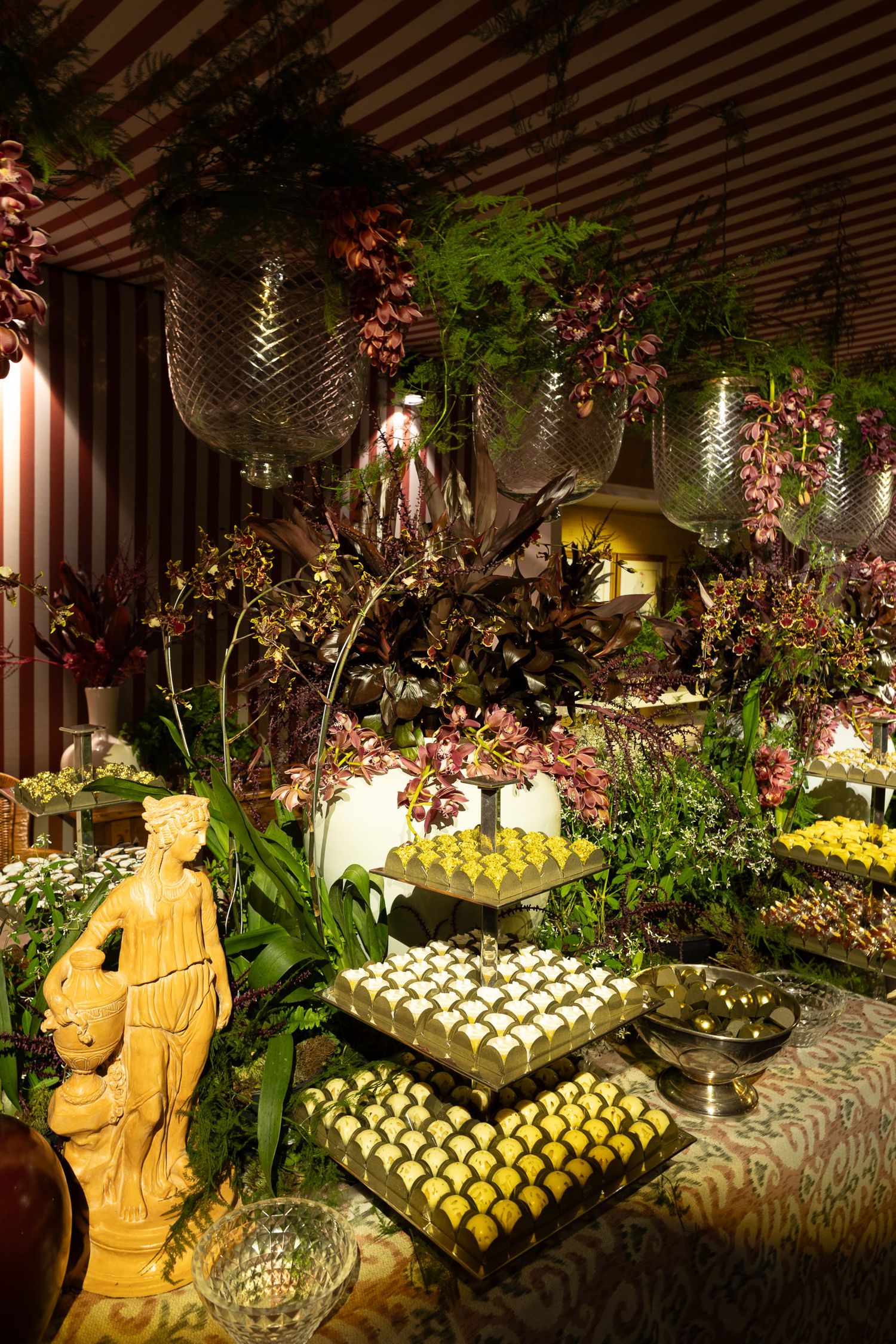 Elegant dessert buffet display with tiered stands of sweets and tropical decor under striped canopy lighting.