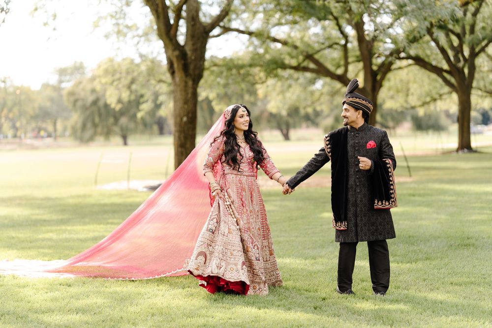 South Asian bride in pink lehenga and groom in black sherwani hold hands walking through green park.