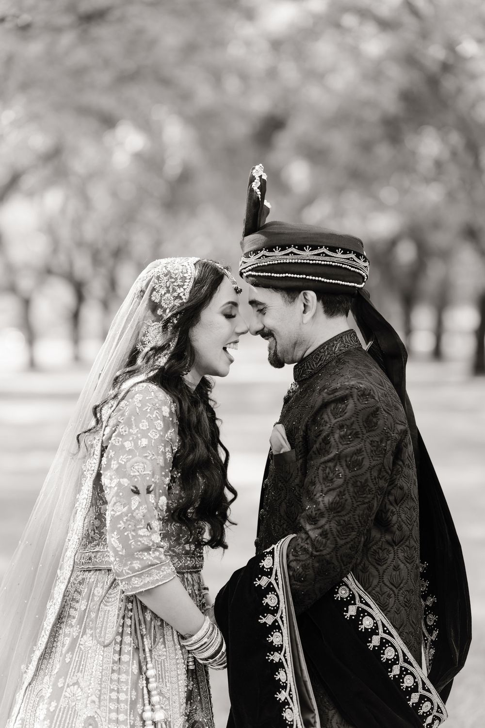 Black and white photo of South Asian wedding couple laughing together in traditional bridal attire outdoors.
