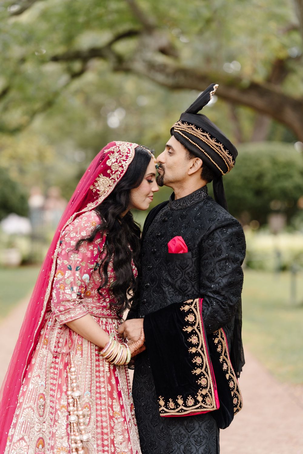 Indian bride in pink lehenga and groom in black embroidered sherwani share romantic moment on garden path.