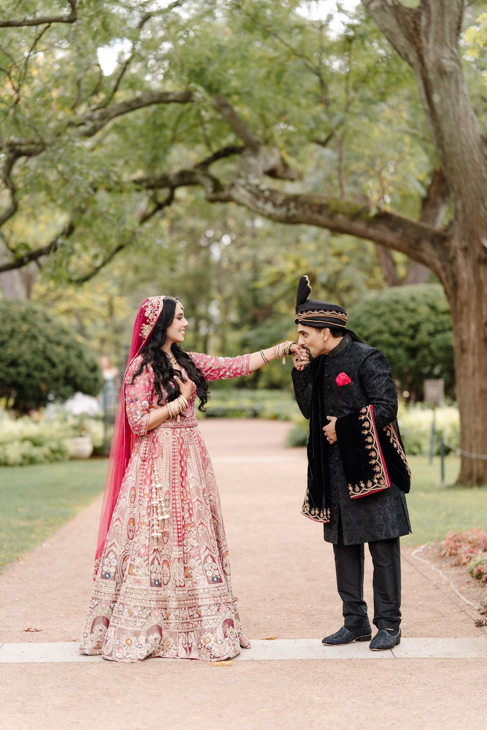 Indian groom kisses bride's hand during outdoor wedding portraits, both wearing traditional South Asian attire.