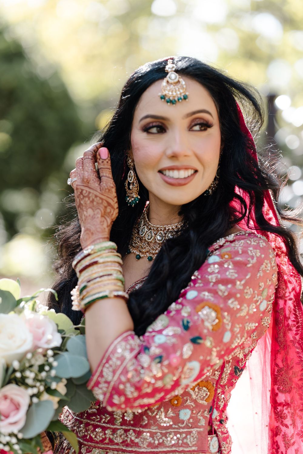Beautiful South Asian bride smiling in pink lehenga with gold jewelry, maang tikka, and mehndi holding bouquet.