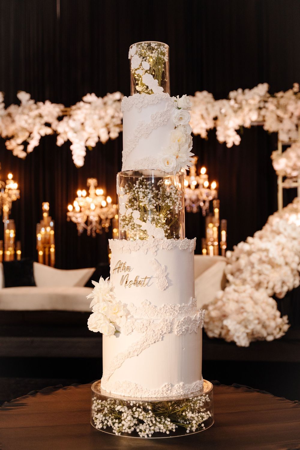 Five-tier white and gold wedding cake with acrylic layers and floral details in candlelit reception venue.