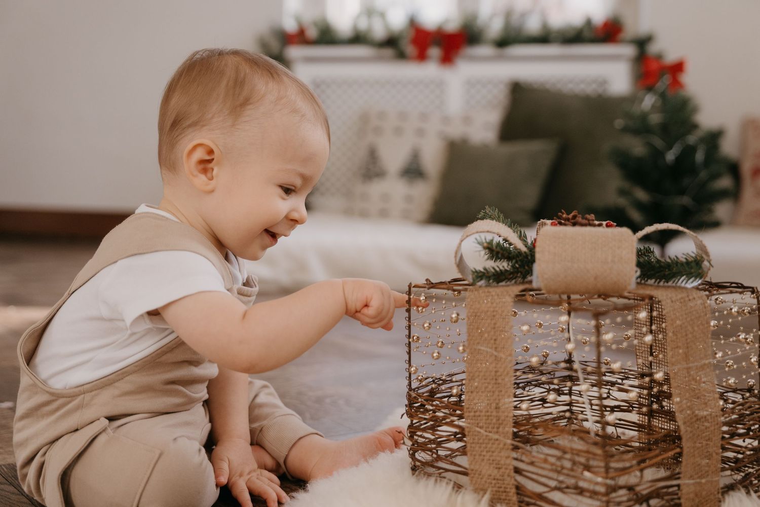 A baby in white and beige clothing reaches for a wrapped Christmas present near a decorated tree.