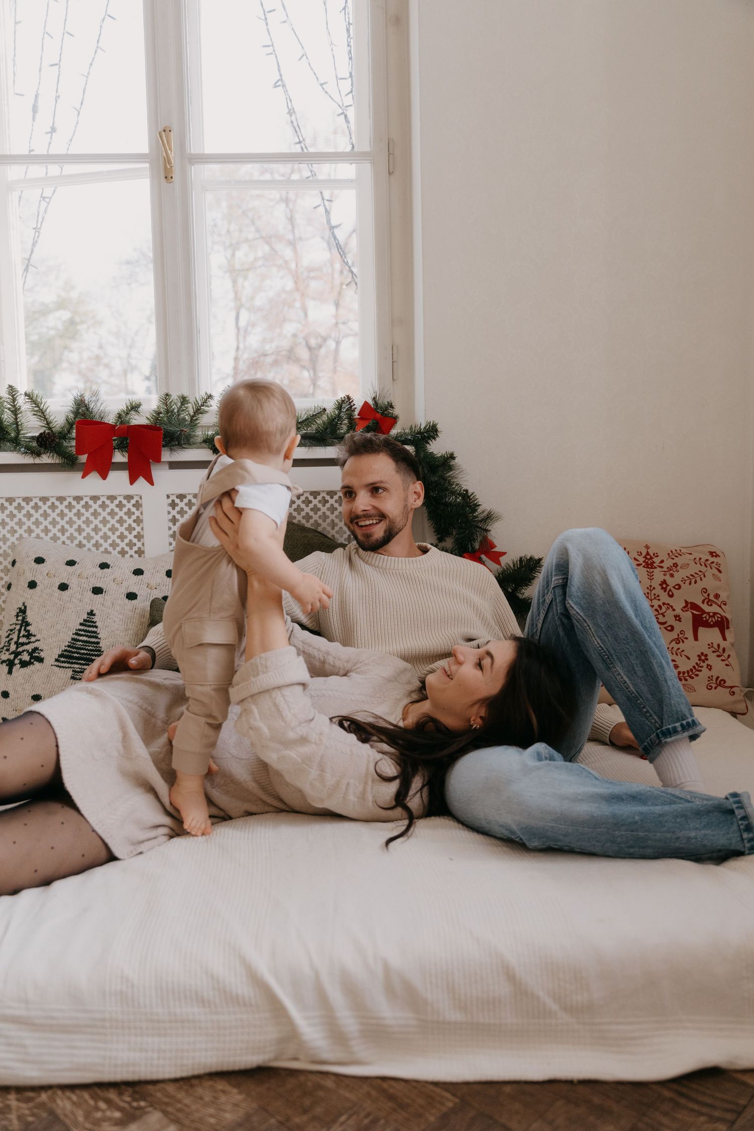 A family relaxes together on a white bed near a window with holiday decorations visible in the background.