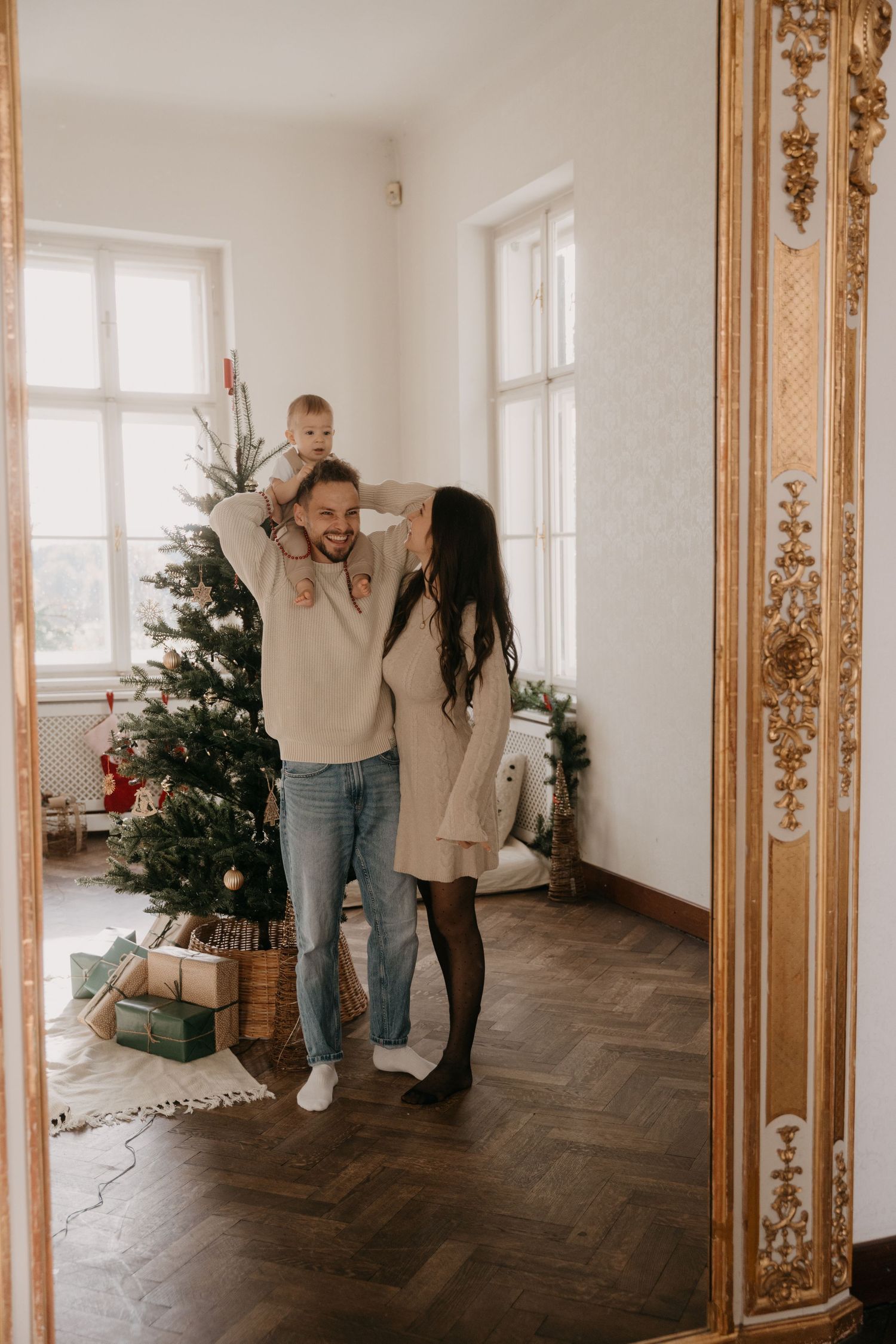 A cozy holiday scene in a bright room with a decorated Christmas tree, herringbone floors and ornate gold trim.