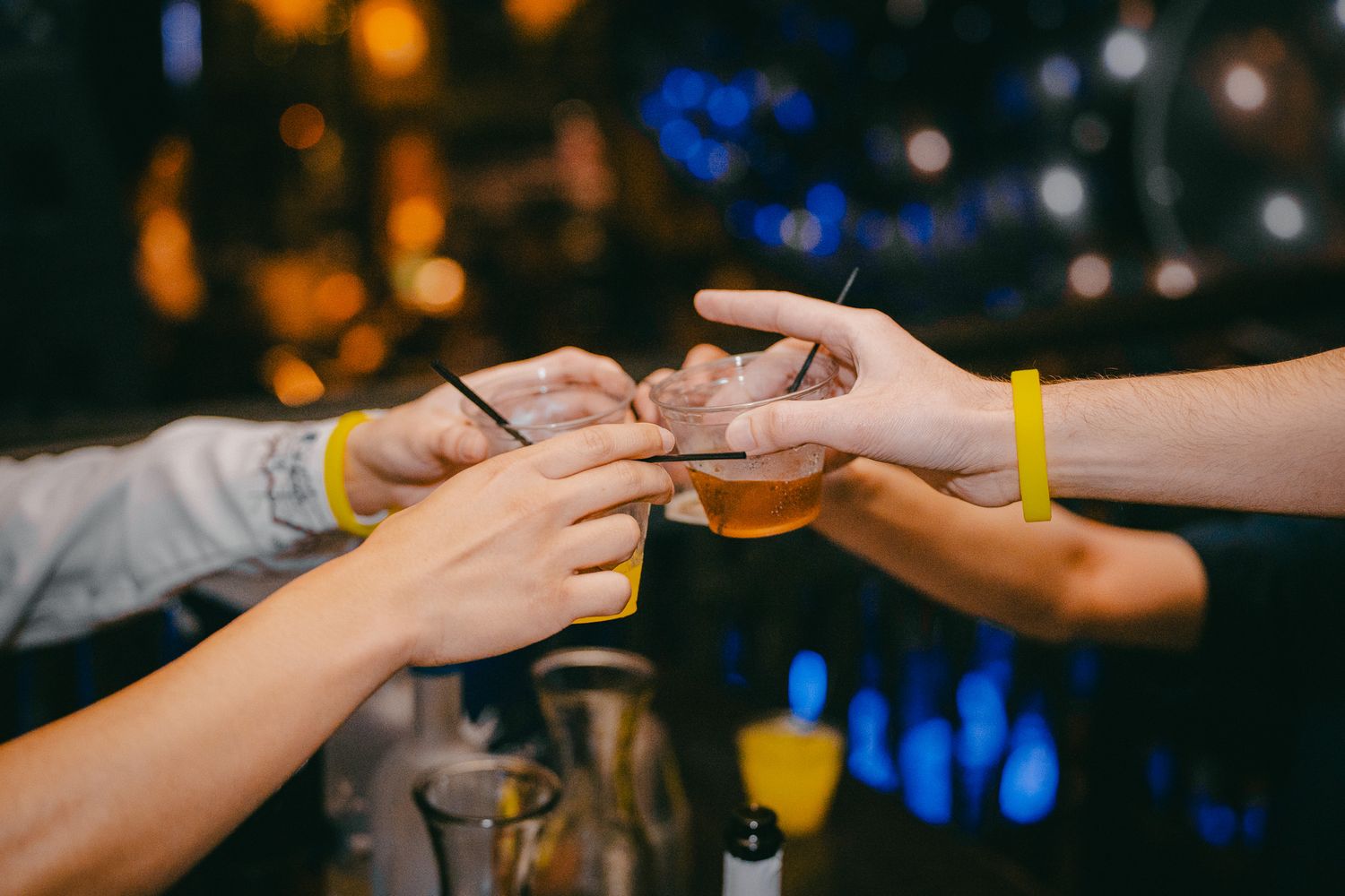 Hands exchanging drinks at a party with colorful bokeh lights in the background.