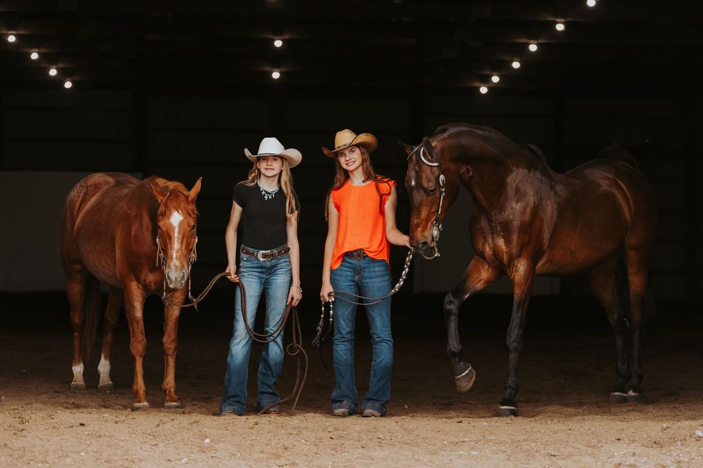 Two cowgirls in hats stand with chestnut horses in an indoor riding arena with string lights overhead.
