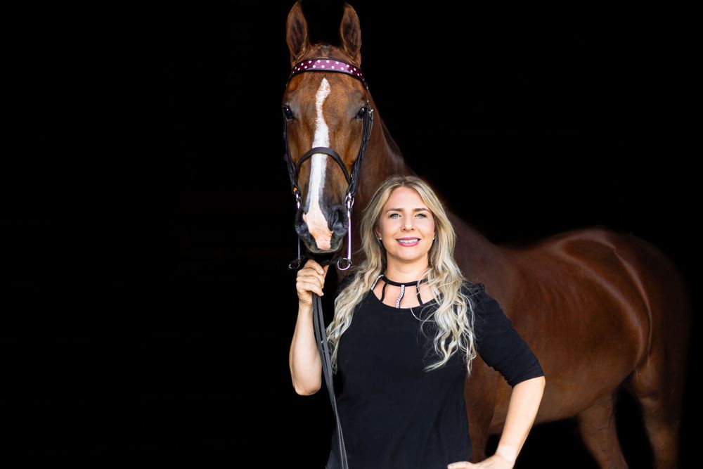 A smiling person poses with a beautiful brown horse against a dark background.