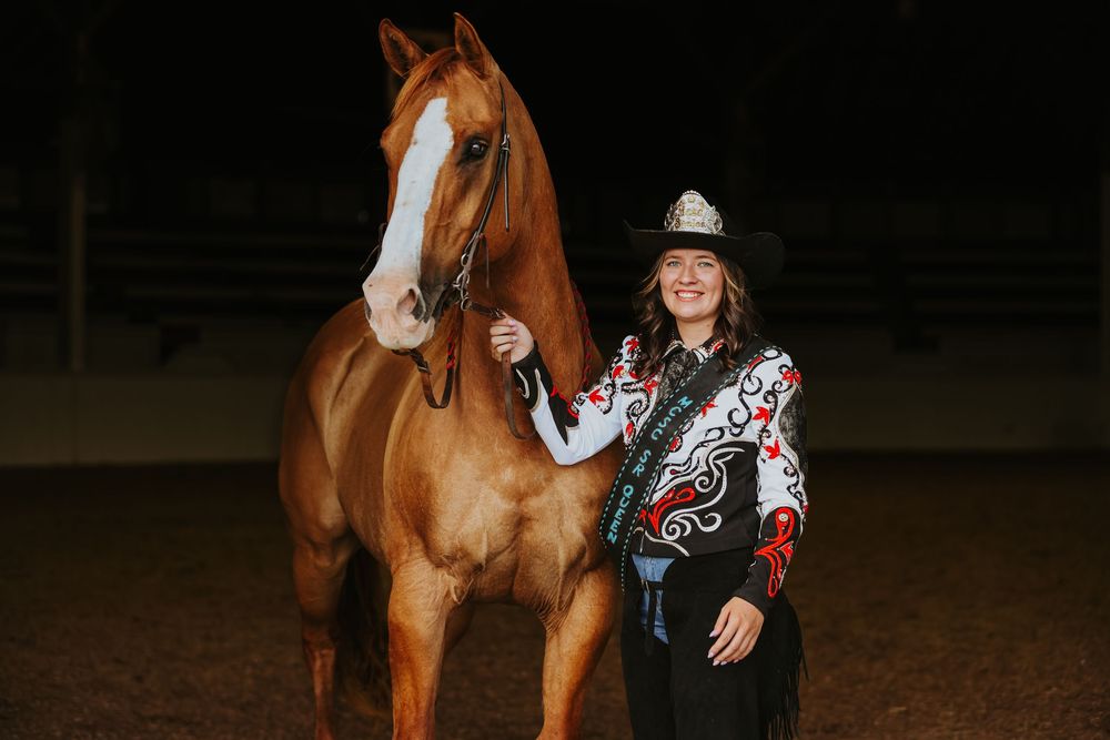 Young woman in western show attire stands proudly beside a chestnut horse in an indoor arena at night.