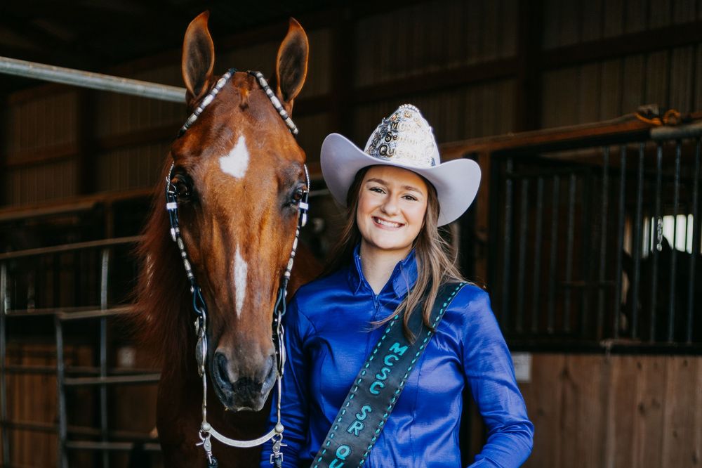 Equestrian in blue jacket and cowboy hat poses with chestnut horse in rustic barn setting.
