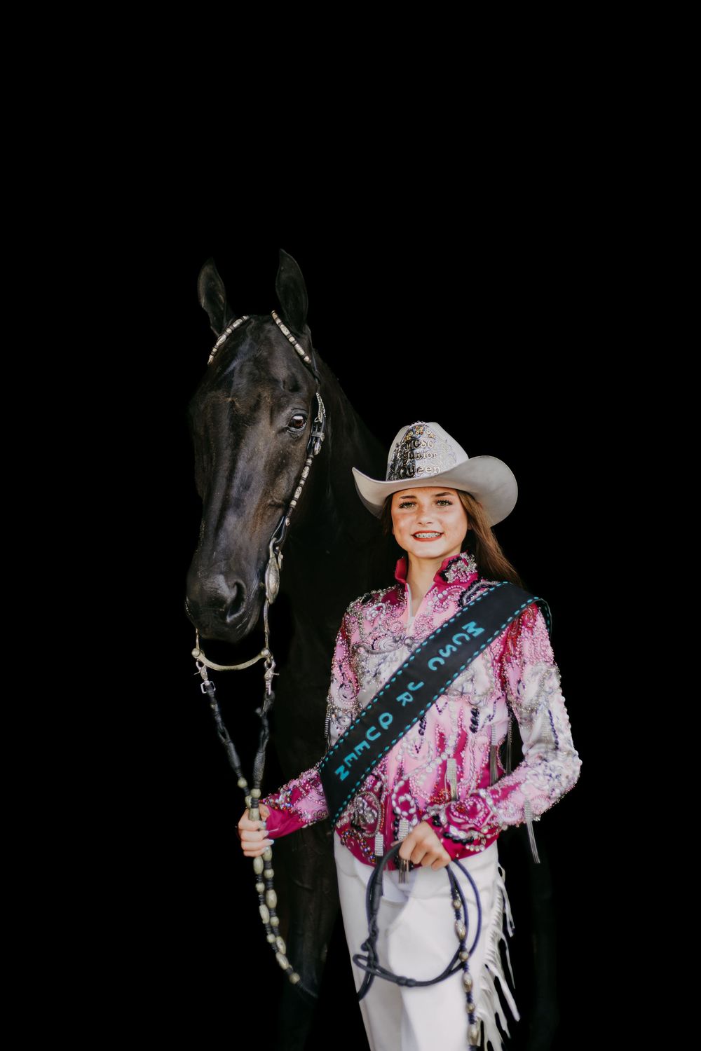 Equestrian poses with dark horse while wearing pink and white show outfit with cowboy hat against black backdrop.