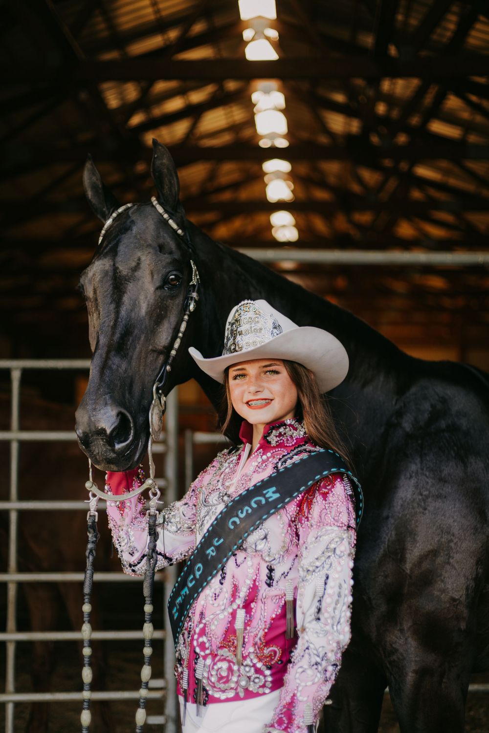 A person in a pink sequined outfit poses with a black horse in a dimly lit indoor riding arena.