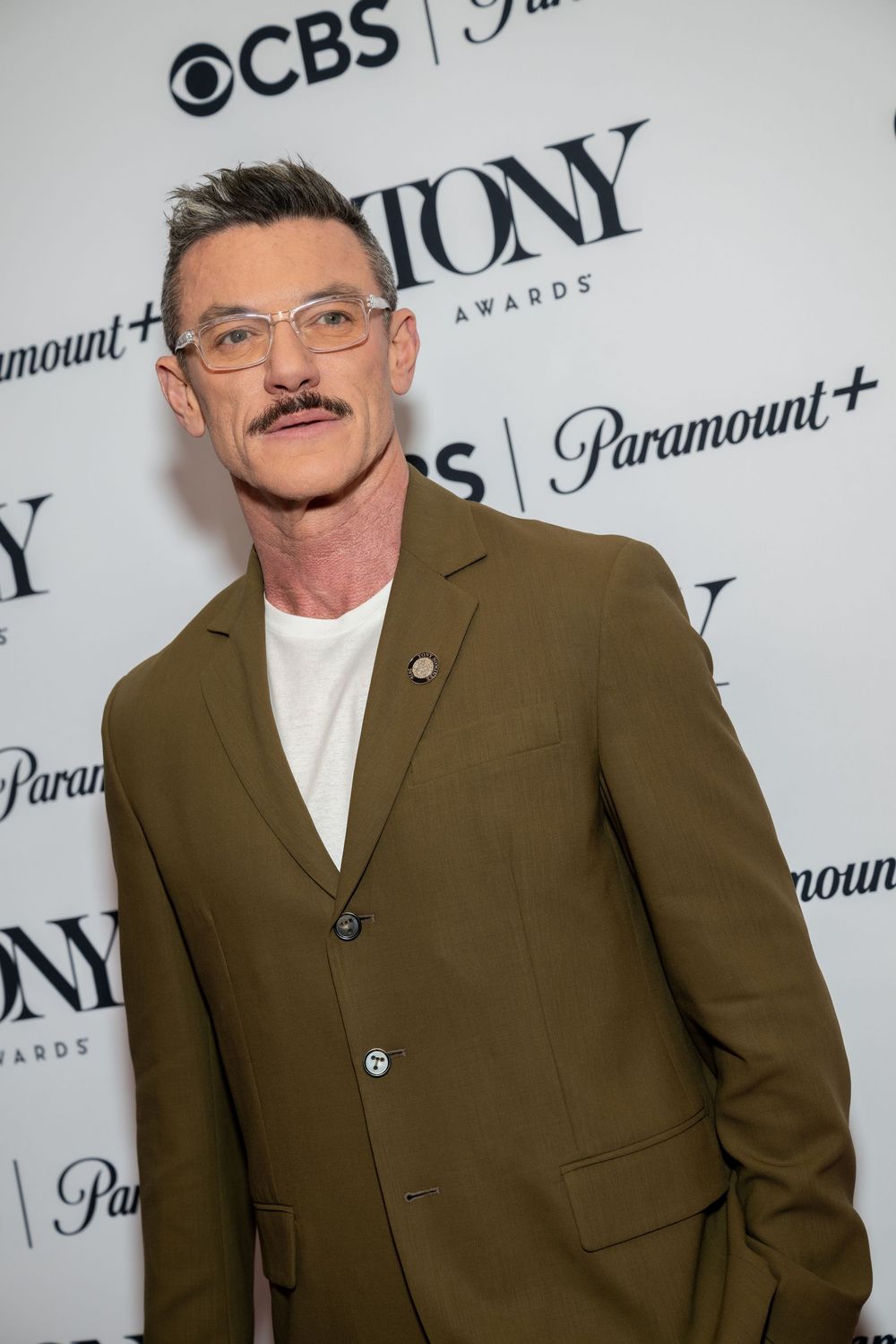 Man in olive blazer poses at Tony Awards CBS Paramount+ event on white step-and-repeat backdrop.