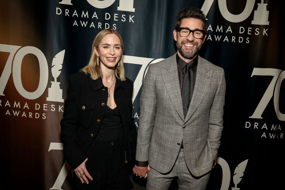 Couple posing together at the 70th Drama Desk Awards, woman in black outfit and man in gray suit.