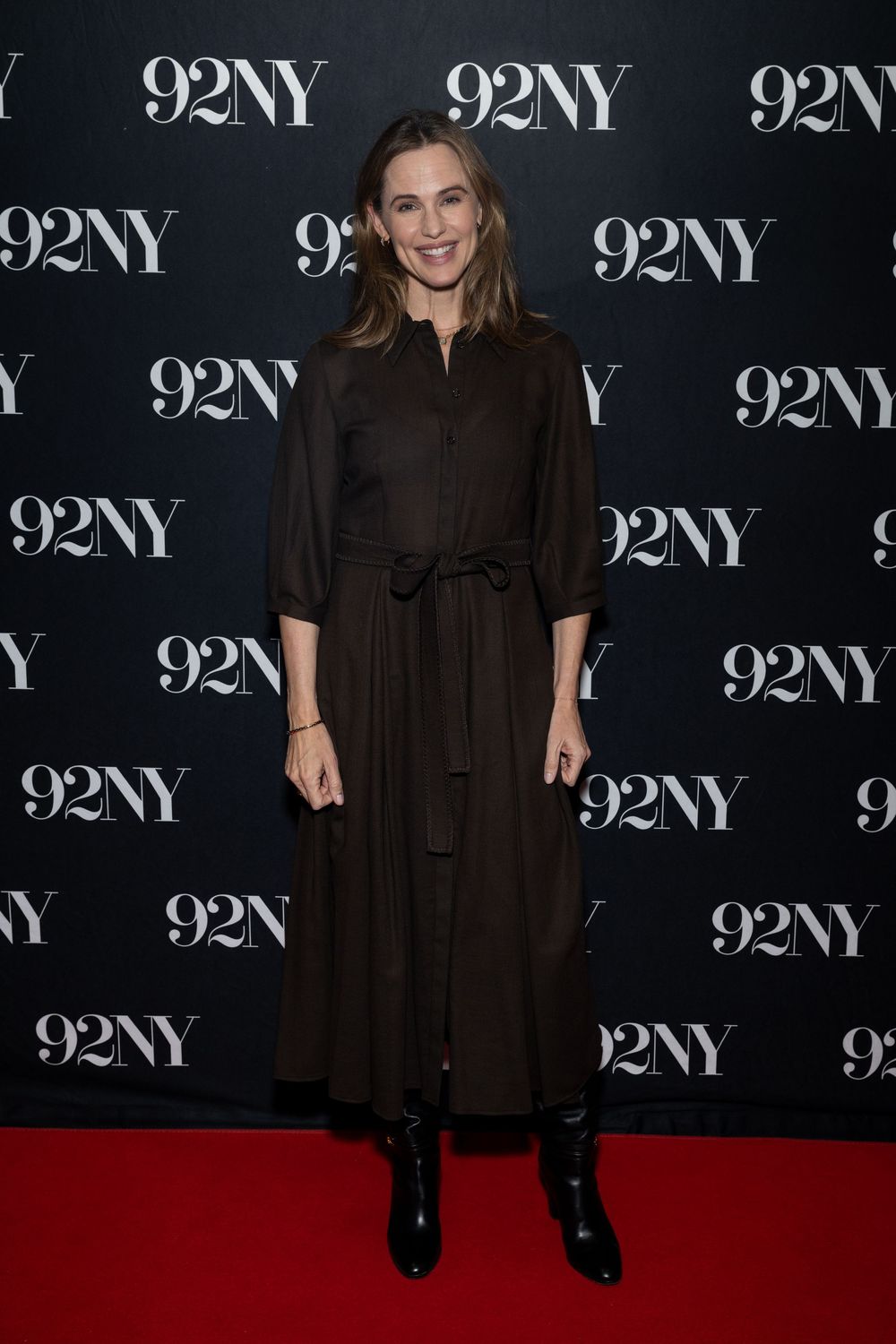 Someone wearing a black dress poses on a red carpet against a backdrop with repeating 92NY logos.