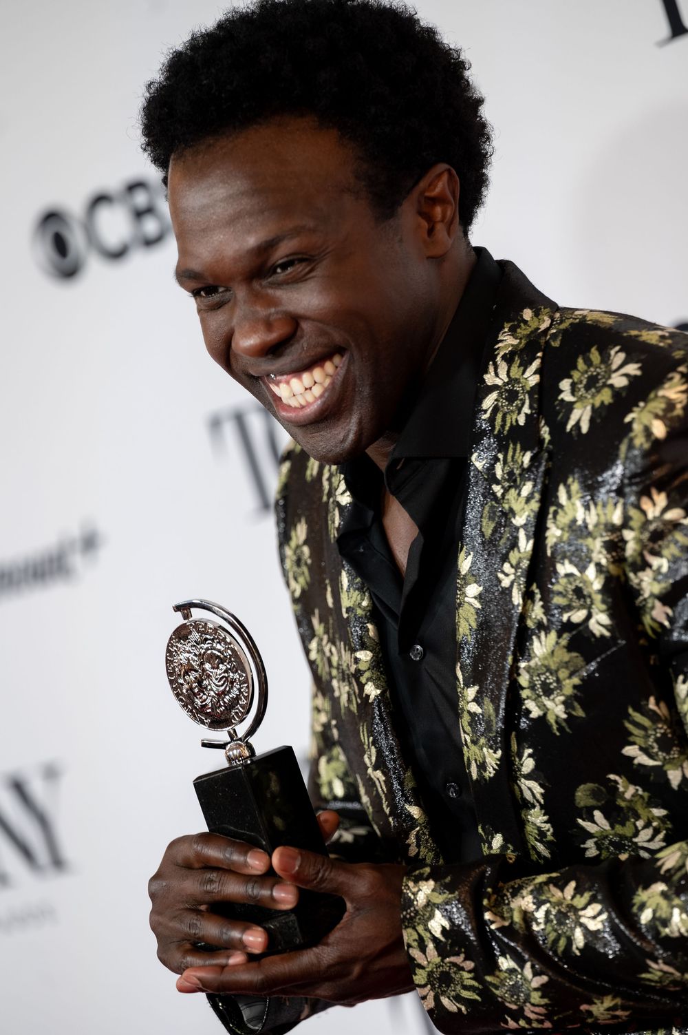 Joyful man in floral gold blazer holding a Tony Award trophy at the CBS Tony Awards press room.