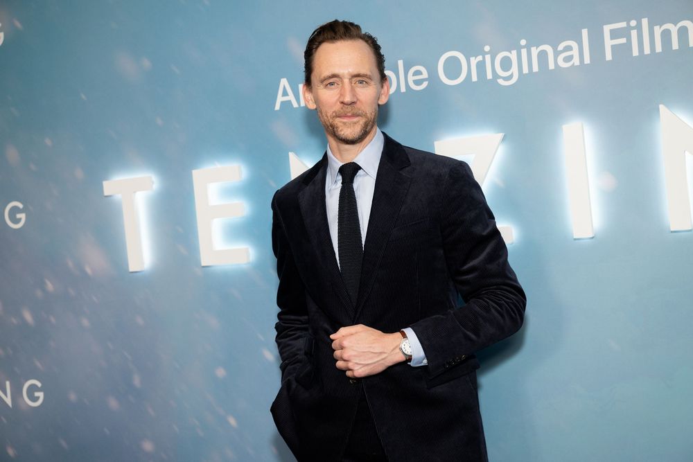 Man in dark suit poses at Amazon Original Films Tenzing movie premiere event on blue backdrop.