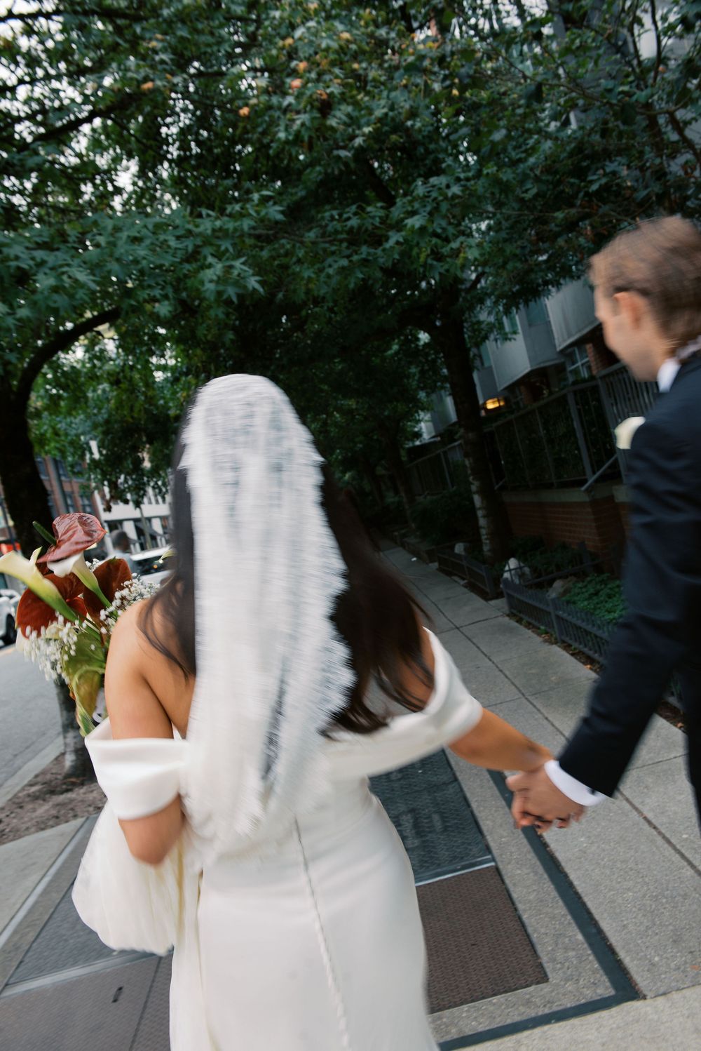 A couple in wedding attire playfully dances on a tree-lined sidewalk.