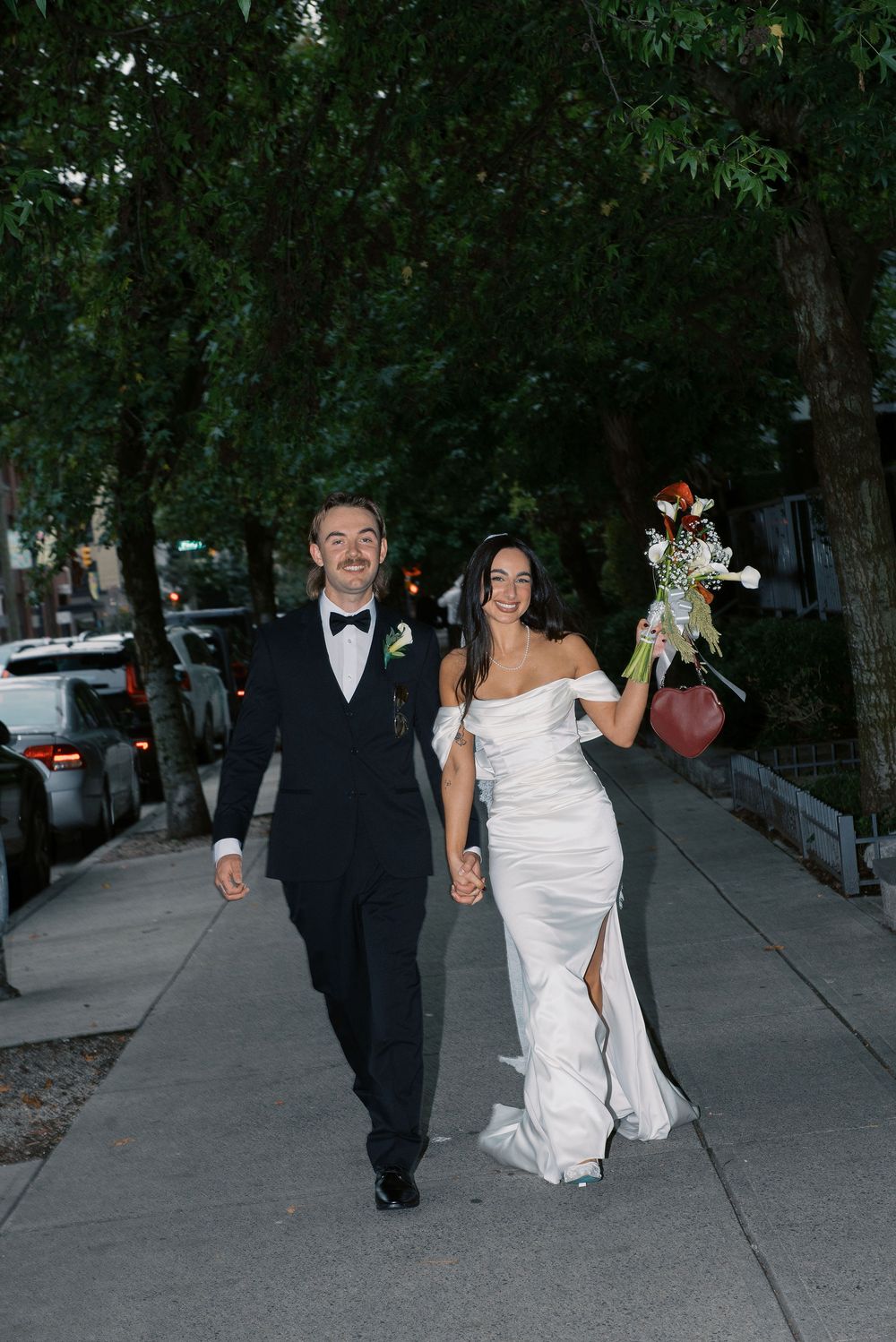 Wedding couple walks down a tree-shaded sidewalk with parked cars visible in the background.