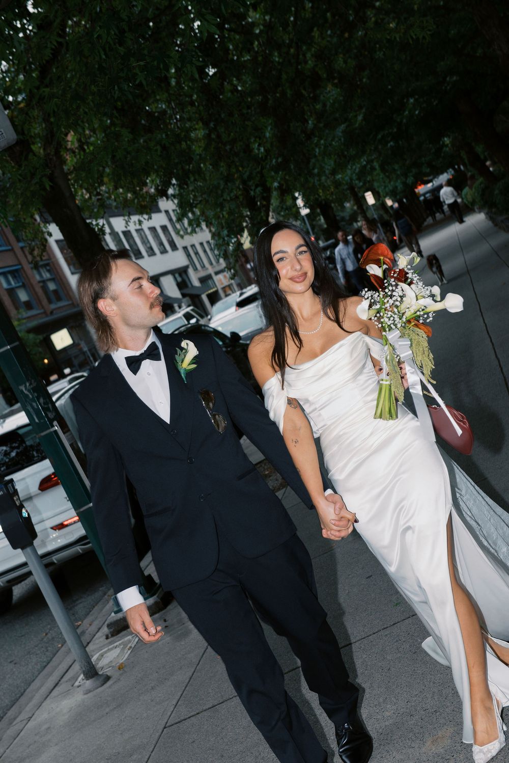 Newlyweds strolling down an urban street with the bride holding a white floral bouquet.
