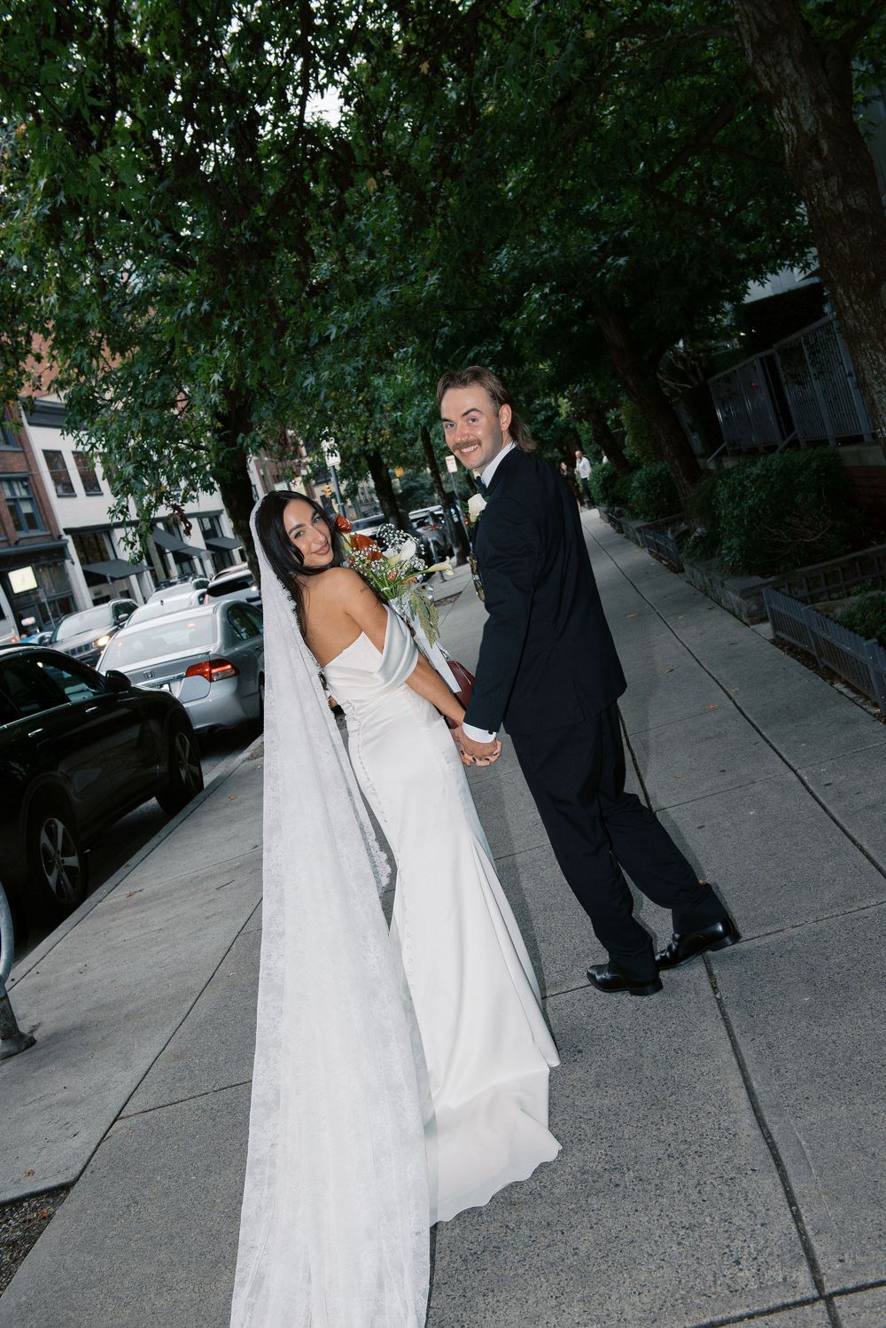 A bride in a flowing white gown and groom in black suit stroll along an urban street.