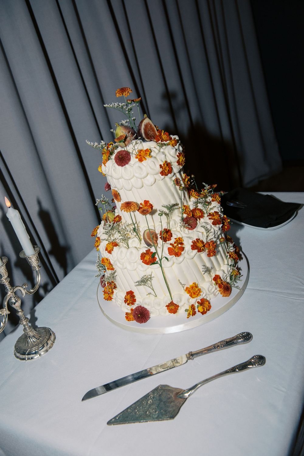 A white wedding cake decorated with fresh figs and orange flowers sits on a table with serving utensils.