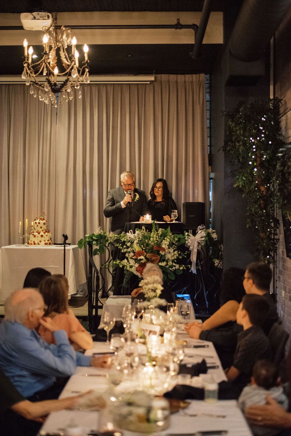 An elegant dinner reception with guests seated at a long table under a crystal chandelier while speakers present at the front.
