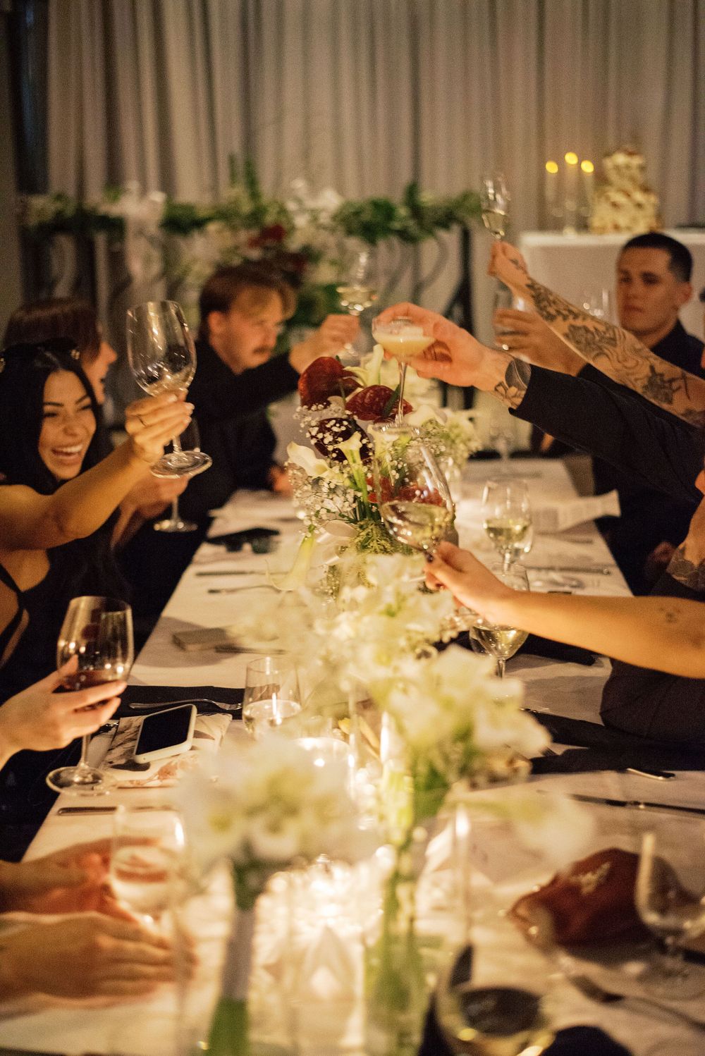 People raising wine glasses in a toast at an elegant dinner party with candlelit table and floral centerpieces.