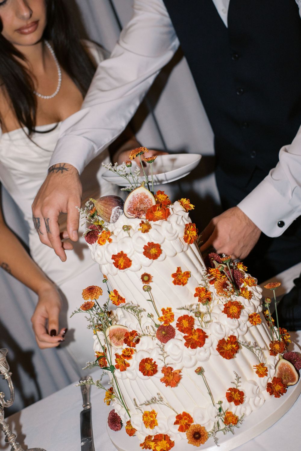 Multiple hands reach to cut slices of an elegant white wedding cake decorated with orange marigold flowers.